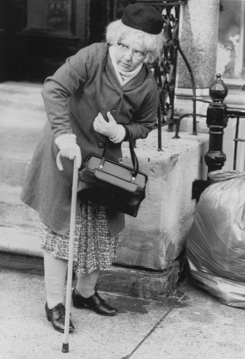 Patricia Moore in disguise, leaning on a cane by a stoop